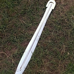 May include: A long, double-edged sword with a silver blade and a gold-colored hilt. The sword lies on a bed of green grass. The blade has etched lines, and the hilt has a rounded pommel.
