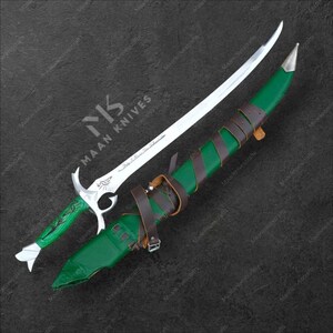 May include: A silver and green fantasy sword with a curved blade, resting on a green sheath with brown leather straps. The handle is green with silver accents. The blade is silver with etched designs. The sheath is green with brown leather straps.