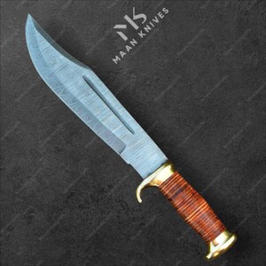Può includere: Un grande coltello in acciaio Damasco con una lama curva. L'impugnatura è avvolta in pelle marrone e presenta dettagli in ottone. Il coltello è posto su uno sfondo scuro. Il testo "MAAN KNIVES" è visibile.