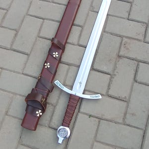 May include: A medieval-style sword with a silver blade and a brown leather-wrapped handle. The sword is next to a brown leather sheath with gold cross accents. The word "VINCT" is visible on the sheath.