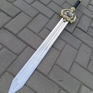 Puede incluir: Una espada de fantasía plateada y dorada con una empuñadura detallada y un agarre negro. La hoja tiene un diseño acanalado. La espada está sobre una superficie de ladrillos grises.