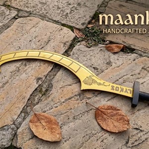 Könnte beinhalten: Ein goldfarbenes, sichelförmiges Schwert mit schwarzem Griff, das eine eingravierte Grafik einer Figur und den Text LYKOS auf der Klinge aufweist. Der Text maanknives HANDCRAFTED ANCIENT BLADES ist in der oberen rechten Ecke zu sehen.