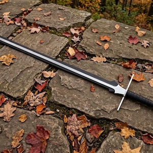 May include: A long sword featuring a black blade with a silver edge, a straight silver crossguard, and a black wrapped handle with a silver pommel. The sword is positioned diagonally across a surface of flat, gray stones covered in fallen autumn leaves.