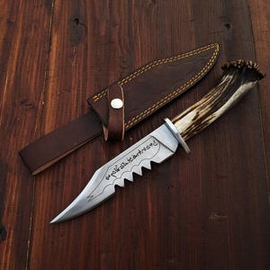 May include: A hunting knife with a serrated blade and a handle made from an antler. The knife is accompanied by a brown leather sheath with a button closure. The blade has an inscription. The knife and sheath are on a dark wooden surface.