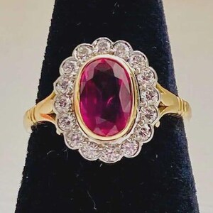 Op de afbeelding: Een gouden ring met een grote, ovaal geslepen dieprode edelsteen in het midden, omringd door een geschulpte halo van kleine, heldere, rond geslepen witte stenen. De ringband is gemaakt van gepolijst geelgoud en vormt een contrast met de rode steen.