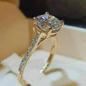 May include: A gold engagement ring featuring a large, clear, round-cut diamond. The band is embellished with smaller diamonds. The ring is presented against a soft, neutral backdrop, highlighting its sparkle and design.