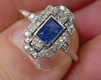 May include: A silver ring featuring a rectangular, deep blue gemstone at the center, surrounded by smaller, clear gemstones. The ring has an Art Deco design with geometric shapes and a delicate band. The gemstones sparkle, creating a luxurious appearance.