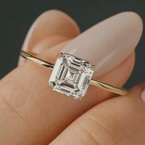 Puede incluir: Un anillo de oro con un gran diamante de talla cuadrada. El diamante es claro y multifacético, reflejando la luz. La banda del anillo es delgada y dorada. El anillo lo sostiene una persona de piel clara.