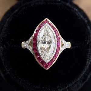 May include: A marquise-cut diamond ring featuring a central elongated stone surrounded by a halo of vibrant red rubies. The setting is crafted from yellow gold with milgrain detailing and small accent diamonds along the split shank band.