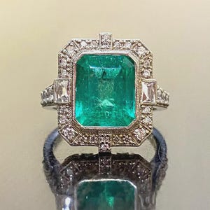 May include: A rectangular emerald-cut emerald ring with a halo of small diamonds. The ring has a vintage design with a rectangular emerald center stone and smaller rectangular stones on the sides. The ring is set in a silver or white gold band.