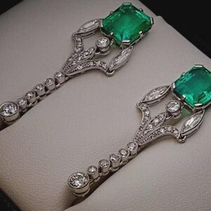 K&ouml;nnte beinhalten: Ein Paar kunstvoller Ohrringe mit quadratisch geschliffenen Smaragden und Diamantbesatz. Die silberfarbenen Ohrringe haben ein dekoratives Design mit einer Reihe kleiner Diamanten, die zu einem gr&ouml;&szlig;eren, runden Diamanten an der Basis f&uuml;hren.
