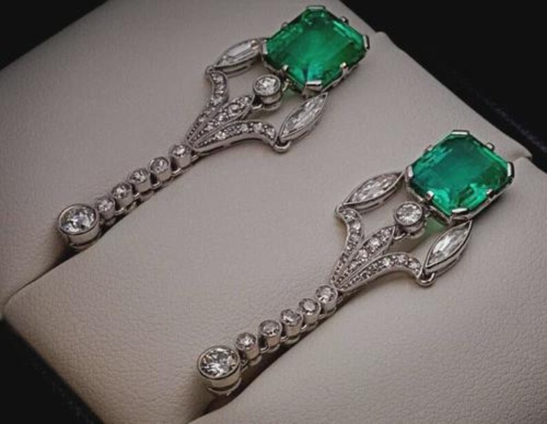 K&ouml;nnte beinhalten: Ein Paar kunstvoller Ohrringe mit quadratisch geschliffenen Smaragden und Diamantbesatz. Die silberfarbenen Ohrringe haben ein dekoratives Design mit einer Reihe kleiner Diamanten, die zu einem gr&ouml;&szlig;eren, runden Diamanten an der Basis f&uuml;hren.
