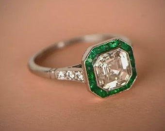 Könnte beinhalten: Ein silberfarbener Ring mit einem zentralen, quadratisch geschliffenen, klaren Stein, der von einer Reihe kleiner grüner Steine umgeben ist. Die Ringschiene ist mit winzigen klaren Steinen besetzt, die in einen glatten Metallschaft übergehen.
