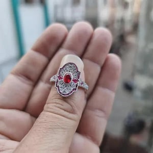 May include: A silver ring featuring a central oval red gemstone, framed by smaller red and clear stones. The ring has an intricate, filigree pattern. The ring is on a finger.