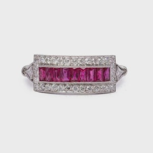 May include: A vintage ring with a rectangular design. The centre features a row of vibrant, rectangular-cut rubies, framed by a border of sparkling, round-cut diamonds. The ring's band is a light silver colour.