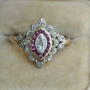 Può includere: Anello in stile vintage con una pietra centrale trasparente di forma ovale, circondata da pietre trasparenti più piccole e un bordo di pietre rosse. L'anello ha una fascia dorata ed è posto su uno sfondo beige.