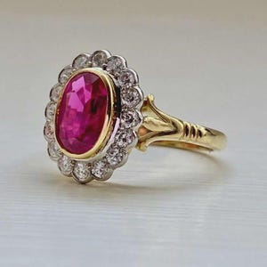 May include: A gold ring featuring a large, oval-cut, vibrant pink gemstone at the center, surrounded by a halo of smaller, clear gemstones. The ring's band is gold, with intricate detailing near the gemstone setting.