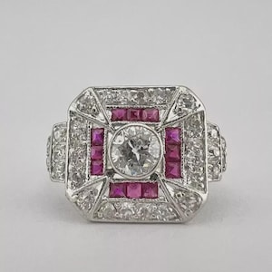 Op de afbeelding: Een platina ring met een centrale ronde diamant, omringd door vierkant geslepen robijnen en kleinere diamanten. De ring heeft een geometrisch ontwerp, met een vierkante vorm en rechthoekige zijaccenten. De ring is geplaatst tegen een effen witte achtergrond.