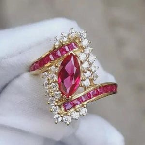 May include: A gold ring featuring a large, marquise-cut ruby center stone, surrounded by smaller diamonds and channel-set rubies. The ring has a unique, asymmetrical design with a cluster of diamonds on one side.