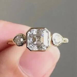 May include: A gold ring featuring a large, square-cut diamond center stone, flanked by two pear-shaped side stones. The ring has a simple, elegant design, showcasing the brilliance of the diamonds. The ring is held up by a person's hand.