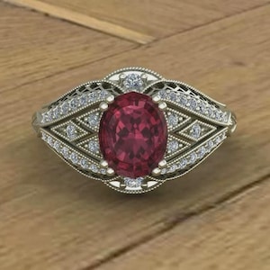 Peut inclure: Une bague en argent ornée d'une grosse pierre précieuse ovale rouge foncé. La bague est ornée de petits diamants transparents et présente un design en filigrane complexe. La bague est posée sur une surface en bois.