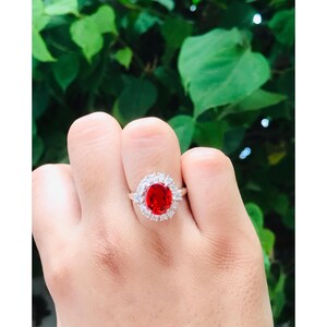 May include: A silver ring featuring a large, oval-cut, bright red gemstone surrounded by a halo of smaller, clear stones. The ring is set against a background of green foliage.