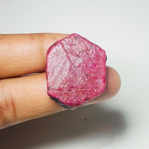 Puede incluir: Una gema de rubí en bruto, hexagonal, de color rosa intenso. La superficie muestra texturas y variaciones naturales, con zonas más oscuras en los bordes. La gema se sostiene entre los dedos sobre un fondo blanco.