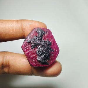 Puede incluir: Una gema de rubí facetada y hexagonal, de color rojo intenso con una inclusión oscura y texturizada. La gema se sostiene entre los dedos, mostrando su apariencia natural y en bruto. La superficie del rubí refleja la luz, resaltando su estructura cristalina.