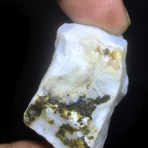 Op de afbeelding: Een ruwe, witte edelsteen met gele plekken en donkergroene insluitsels. De steen wordt tussen vingers gehouden tegen een zwarte achtergrond, wat de natuurlijke, onbewerkte textuur en kleurvariaties benadrukt.