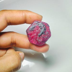 May include: A raw, unpolished ruby gemstone. The ruby is a deep crimson colour with dark grey inclusions. The gemstone is held between fingers, highlighting its natural, irregular shape and texture. The ruby is a precious stone.