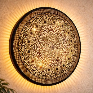 Puede incluir: Aplique de pared redondo de metal negro con un intrincado diseño floral. La lámpara emite una cálida luz dorada, creando un ambiente decorativo y acogedor. La lámpara está montada en una pared beige texturizada.