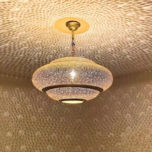 May include: A gold-colored, ornate pendant light fixture with a perforated design. The light is suspended from the ceiling and casts a patterned shadow. The fixture has a layered, rounded shape and a warm, inviting glow.