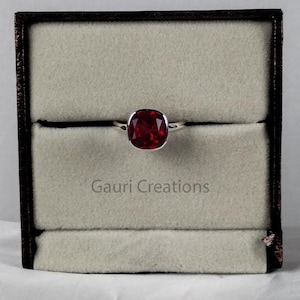 May include: A silver ring with a large, oval, deep red gemstone is displayed inside a brown jewelry box with a light beige interior. The text "Gauri Creations" is visible on the box.