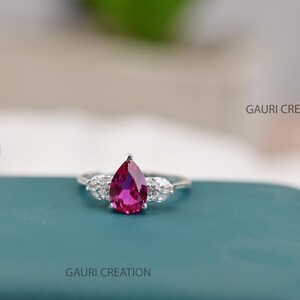 Op de afbeelding: Een zilveren ring met een centrale peervormige, felroze edelsteen, geflankeerd door twee kleinere, heldere, marquise-geslepen stenen aan weerszijden. De tekst GAURI CREATION is zichtbaar, evenals een G-logo in de hoek van de afbeelding.