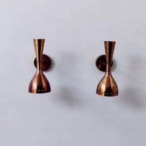 Könnte beinhalten: Zwei kupferfarbene Wandleuchten im Mid-Century-Modern-Design. Jede Leuchte hat einen ausgestellten, sanduhrförmigen Schirm und eine kreisförmige Montageplatte. Auf der linken Seite des Bildes befindet sich ein schwarzes, kreisförmiges Objekt.
