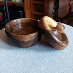 Könnte beinhalten: Eine handgefertigte Holzschale mit Deckel. Schale und Deckel bestehen aus dunkelbraunem und hellbraunem Holz. Der Deckel hat einen kleinen, abgerundeten Stein als Griff. Die Schale steht auf einer hellgrauen Oberfläche.