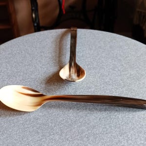 Könnte beinhalten: Zwei Löffel aus Horn mit einem natürlichen Muster aus hellbeige und dunkelbraun. Ein Löffel befindet sich im Vordergrund und der andere im Hintergrund. Die Löffel haben eine glatte, polierte Oberfläche.