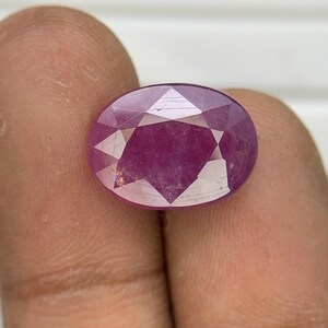 May include: An oval-cut gemstone with a deep magenta-pink hue and a faceted surface. The stone displays a clear, symmetrical pattern of triangular and kite-shaped facets that reflect light across its polished, translucent, and vibrant surface.