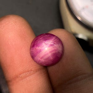 May include: A round, polished gemstone with a deep magenta hue and a star-like effect. The gem has a smooth, reflective surface with internal light patterns. The image is a close-up, highlighting the gem's color and texture. The gem is held between two fingers.