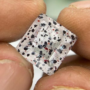 Puede incluir: Una piedra preciosa transparente de corte cuadrado con numerosas inclusiones pequeñas de color negro oscuro y rojo vibrante dispersas por su estructura cristalina clara, con una superficie facetada que refleja la luz en su forma.