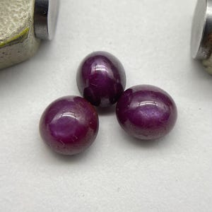 May include: Three polished, deep red gemstones with a glossy finish. The stones are round and appear to be cabochons. The background is a light, neutral color, and the gemstones are the focal point.