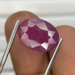 Pode incluir: Uma pedra preciosa de cor magenta, translúcida e de formato oval com uma superfície facetada. A pedra apresenta um padrão distinto em forma de estrela esculpido na face superior, que reflete a luz e exibe inclusões naturais internas.