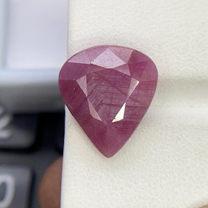 May include: A pear-shaped, faceted gemstone with a deep magenta-pink hue. The stone features a triangular cut pattern on its surface, displaying visible internal inclusions and a polished, reflective finish that catches the light.
