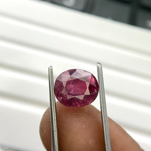 May include: A faceted, oval-shaped, deep red gemstone is held by tweezers. The gem has a brilliant cut, reflecting light. The background includes a calculator and a white surface.