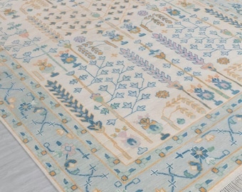 May include: A rectangular area rug with a cream-colored field and a light blue border. The rug features a repeating floral pattern in shades of blue, green, pink, and tan. The border has a geometric design with fringe along the edge.