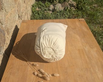 May include: A cream-colored, drawstring bag filled with a fluffy material, likely a pillow or cushion. The bag sits on a light brown wooden surface, with a bundle of dried, beige decorative grass nearby. The background features a stone wall and greenery.