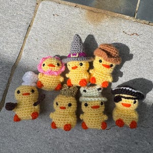 May include: A group of handmade crochet yellow ducklings, each wearing a different hat. Hats include a chef's hat, a witch's hat, and a crown. The ducklings have orange feet and beaks.