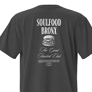 Könnte beinhalten: Dunkelgraues T-Shirt mit weißem Aufdruck auf dem Rücken. Der Aufdruck enthält den Text "SOULFOOD BRONX", eine Burger-Illustration und den Text "The Good Toasted Club". Das T-Shirt hat kurze Ärmel und einen Rundhalsausschnitt.