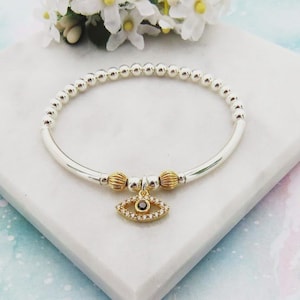May include: Silver beaded bracelet with a gold evil eye charm. The charm is encrusted with small, clear crystals and a black centre. The bracelet also features gold accent beads. The bracelet is displayed on a white marble surface.