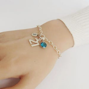 May include: A silver chain link bracelet featuring three charms: a small circular disc engraved with the letter S, a large numeral 21, and a round, faceted blue gemstone set in a silver bezel. The bracelet is secured with a silver clasp.